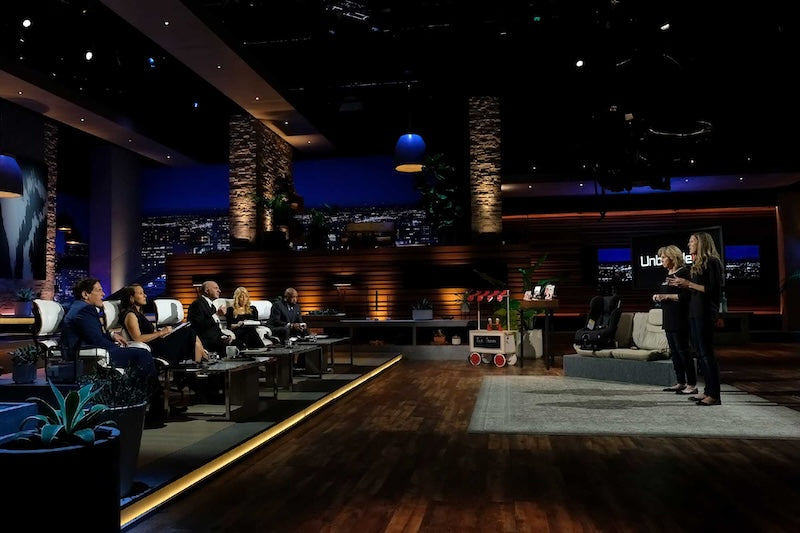 Shark Tank TV show studio set where people are standing and talking with a cityscape background.