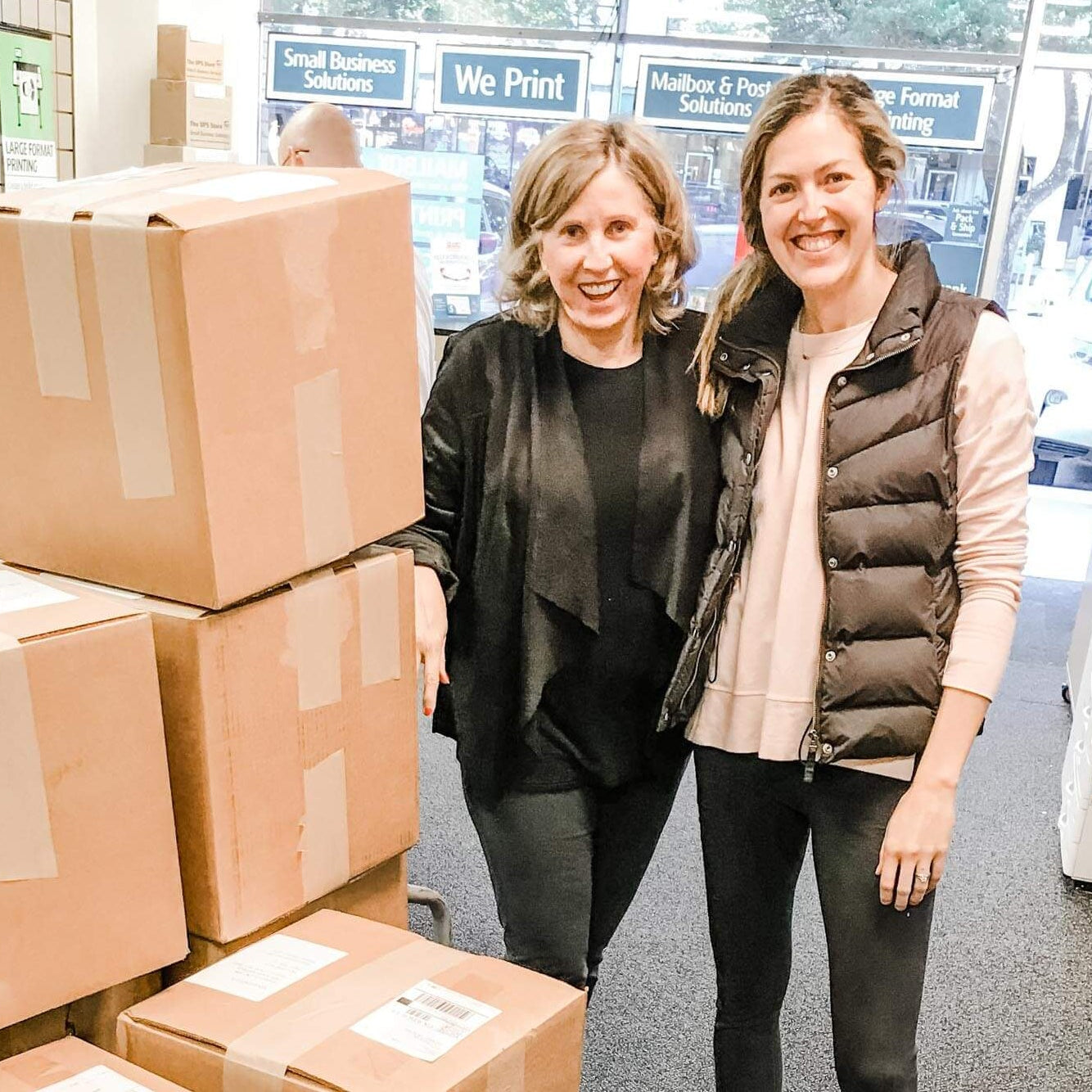 Two women standing next to a stack of cardboard boxes in a printing or packaging facility.